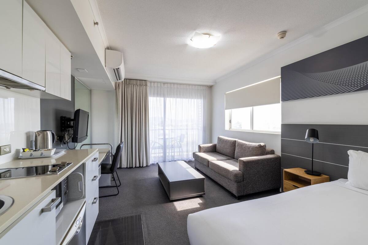 A modern hotel room with a kitchenette, a sofa, a bed, and a large window.
