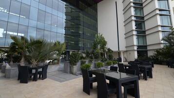 Un patio exterior con mesas y sillas, rodeado de plantas en macetas y un edificio moderno con ventanas de vidrio reflectante.