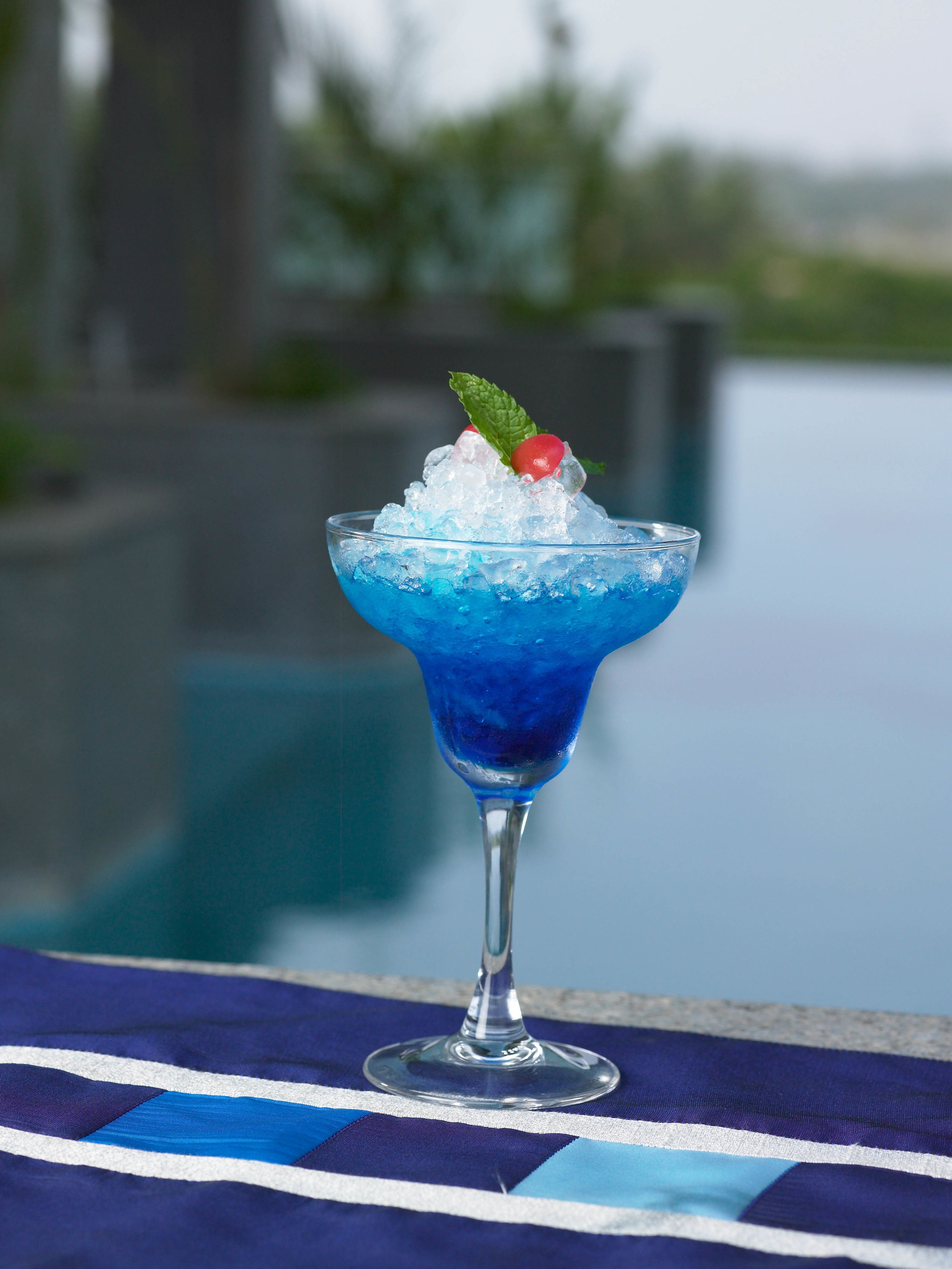 A blue cocktail in a glass with ice and garnishes, placed on a striped cloth.