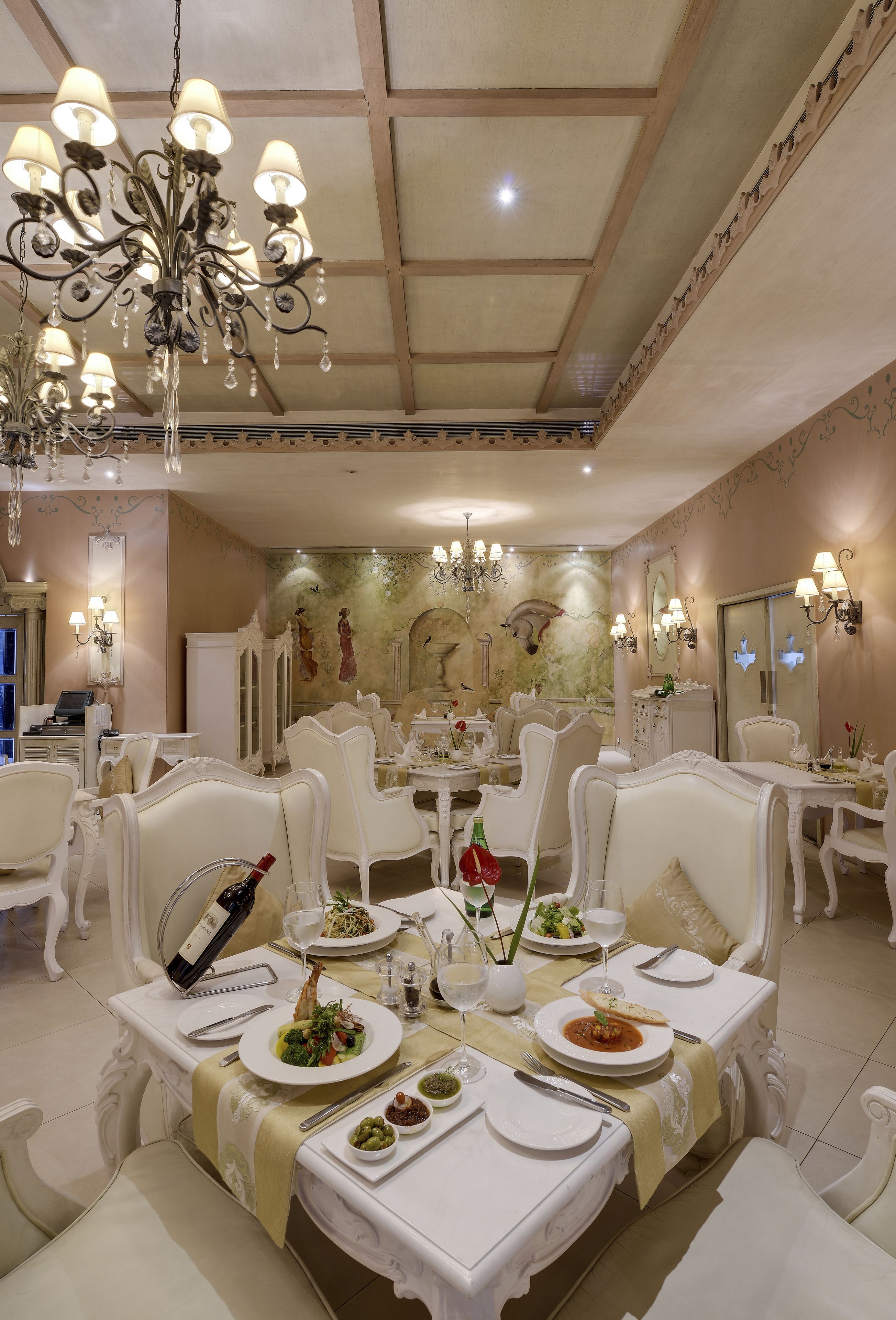 A dining area with white ornate furniture, chandeliers, and a mural on the wall.