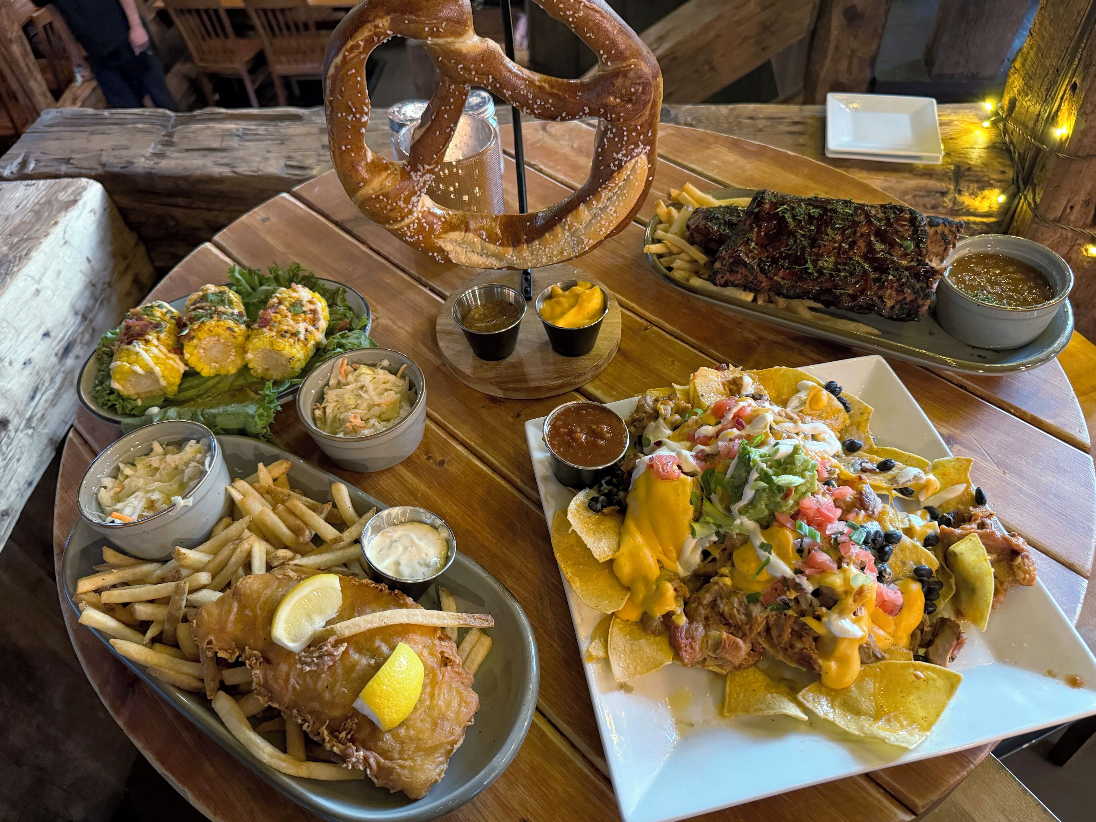 A variety of dishes including fish, fries, nachos, and a pretzel on a wooden table.