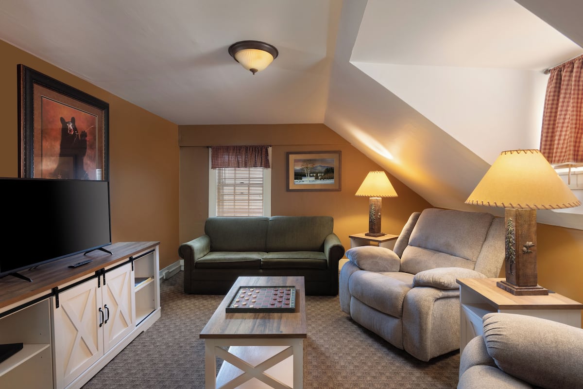 A cozy living room with a flat-screen TV, a green sofa, a grey armchair, a coffee table, and a lamp.