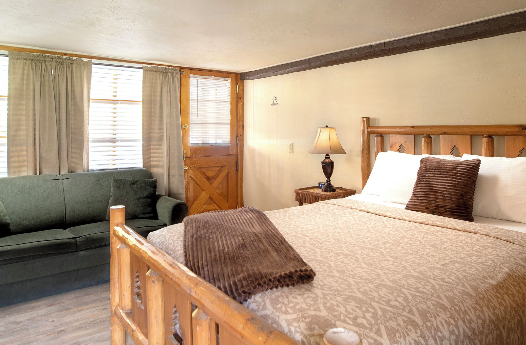 A room with a bed, a wooden headboard, a bedside table with a lamp, a green sofa, and a window with curtains.