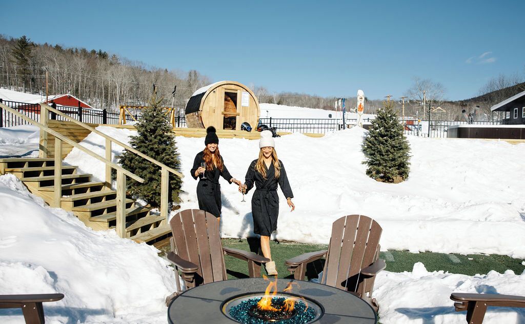 A snowy outdoor area with a wooden deck, a fire pit table, and two Adirondack chairs.