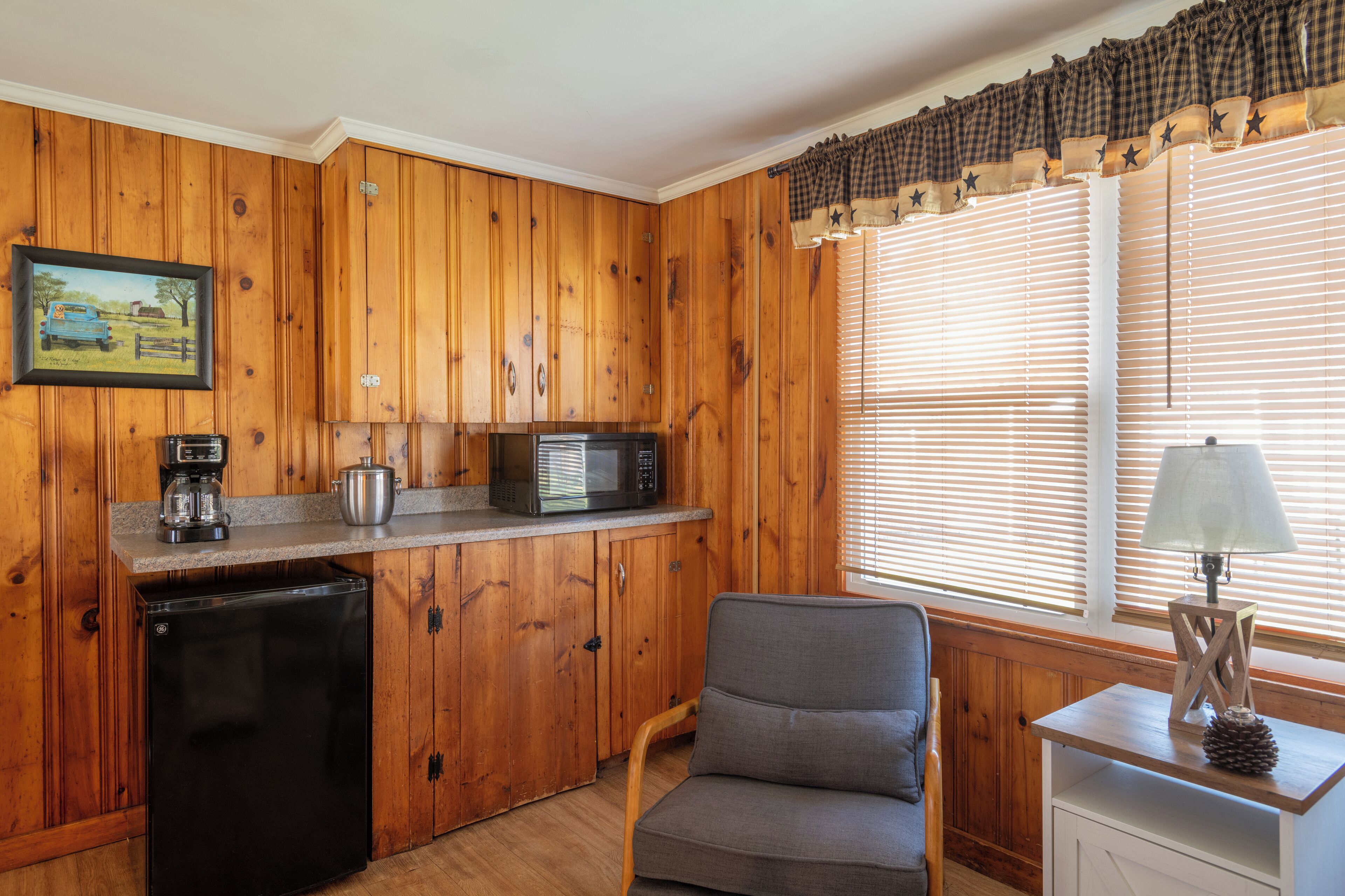 Cocina compacta con paneles de madera, un refrigerador, un microondas, una cafetera y una ventana con persianas.