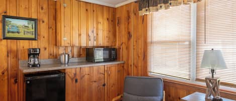 A compact kitchen with wooden paneling, a refrigerator, a microwave, a coffee maker, and a window with blinds.