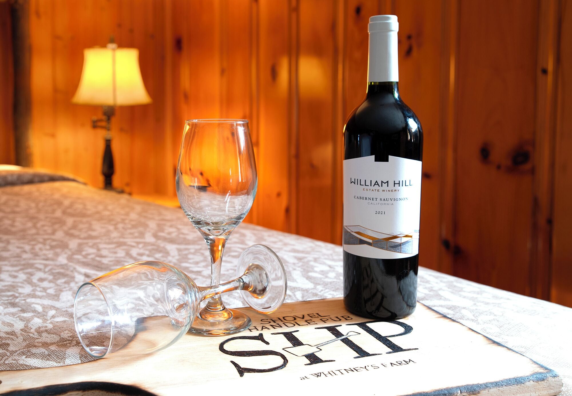 Una botella de vino Cabernet Sauvignon 2021 de la bodega William Hill Estate y una copa sobre una mesa, con un fondo de madera y una lámpara.