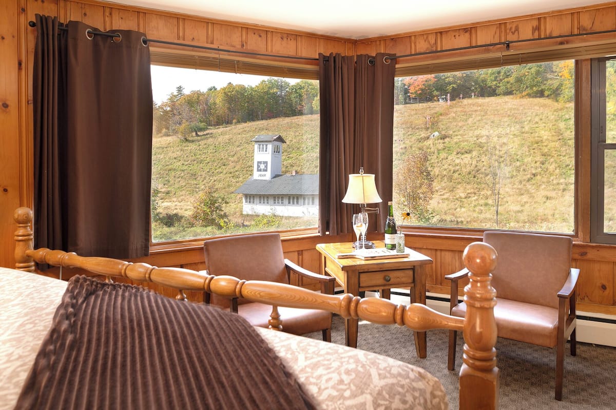 A hotel room with a large bed, two wooden chairs, a small table with a lamp, and a view of a hill with a building.