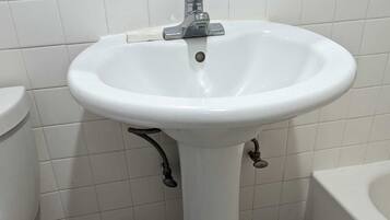 Een badkamer met een wit wastafeltje op voet, twee kraan, een wit toilet, een witte prullenbak en een bad.