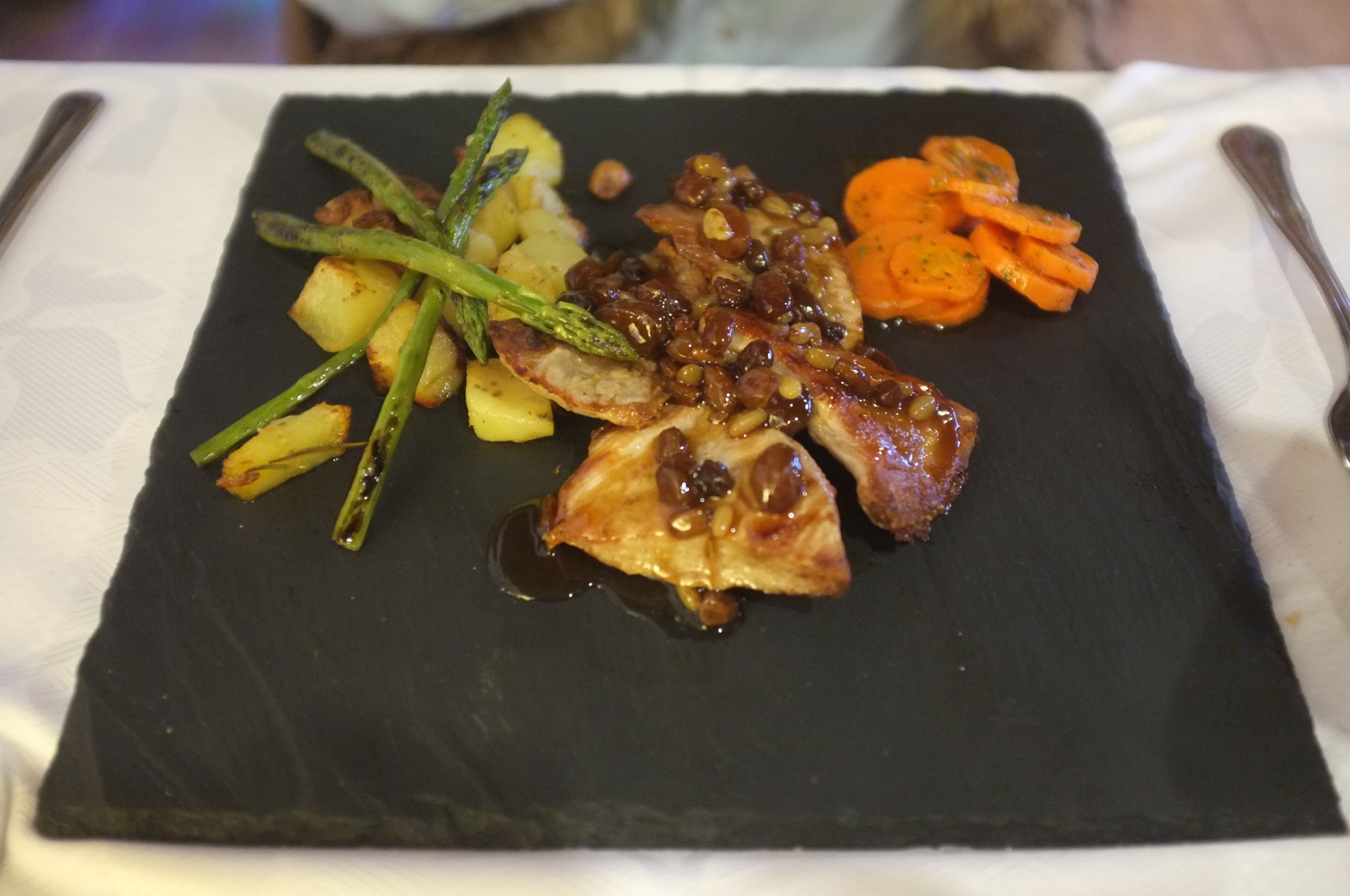 A square black plate with a piece of meat, roasted potatoes, asparagus, and carrots.