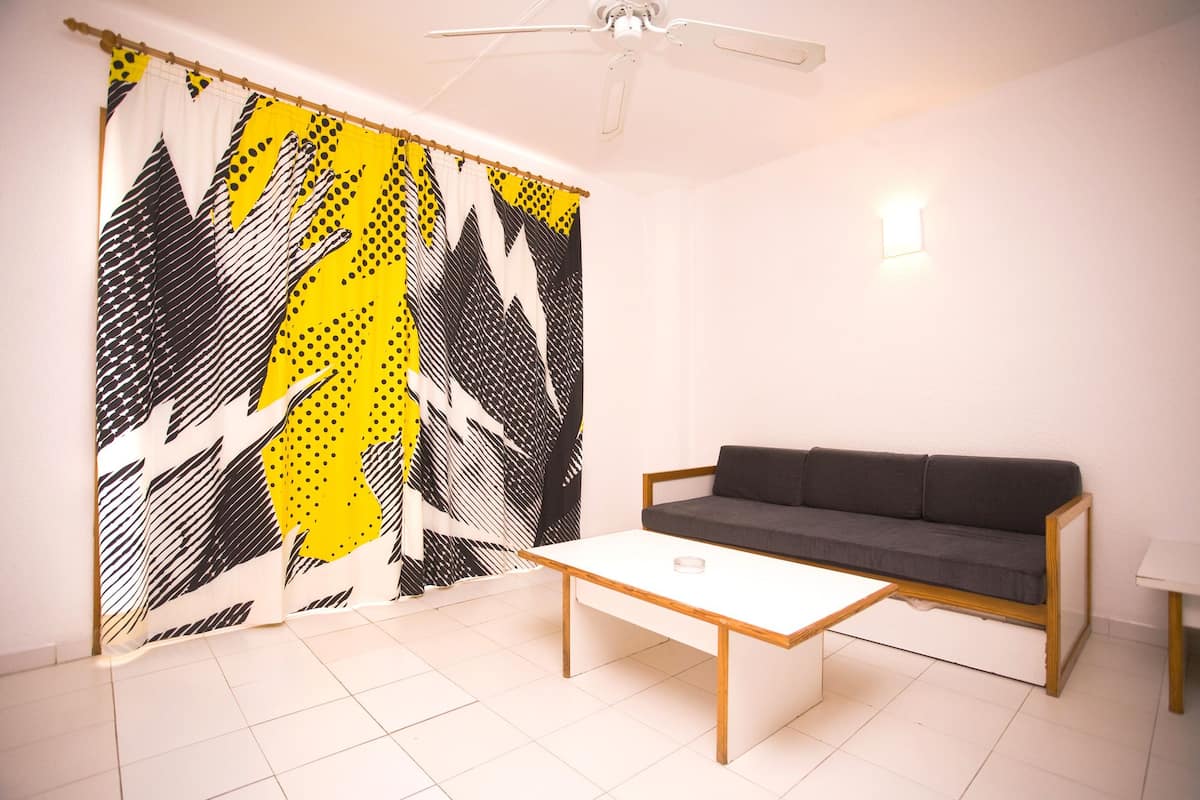 Una habitación con ventilador de techo blanco, sofá gris, mesa de centro blanca y cortina estampada.