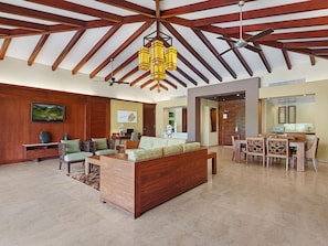 A spacious living area with a high ceiling, wooden beams, a large wooden sofa, a dining table with chairs, and a kitchen area visible through an open doorway.