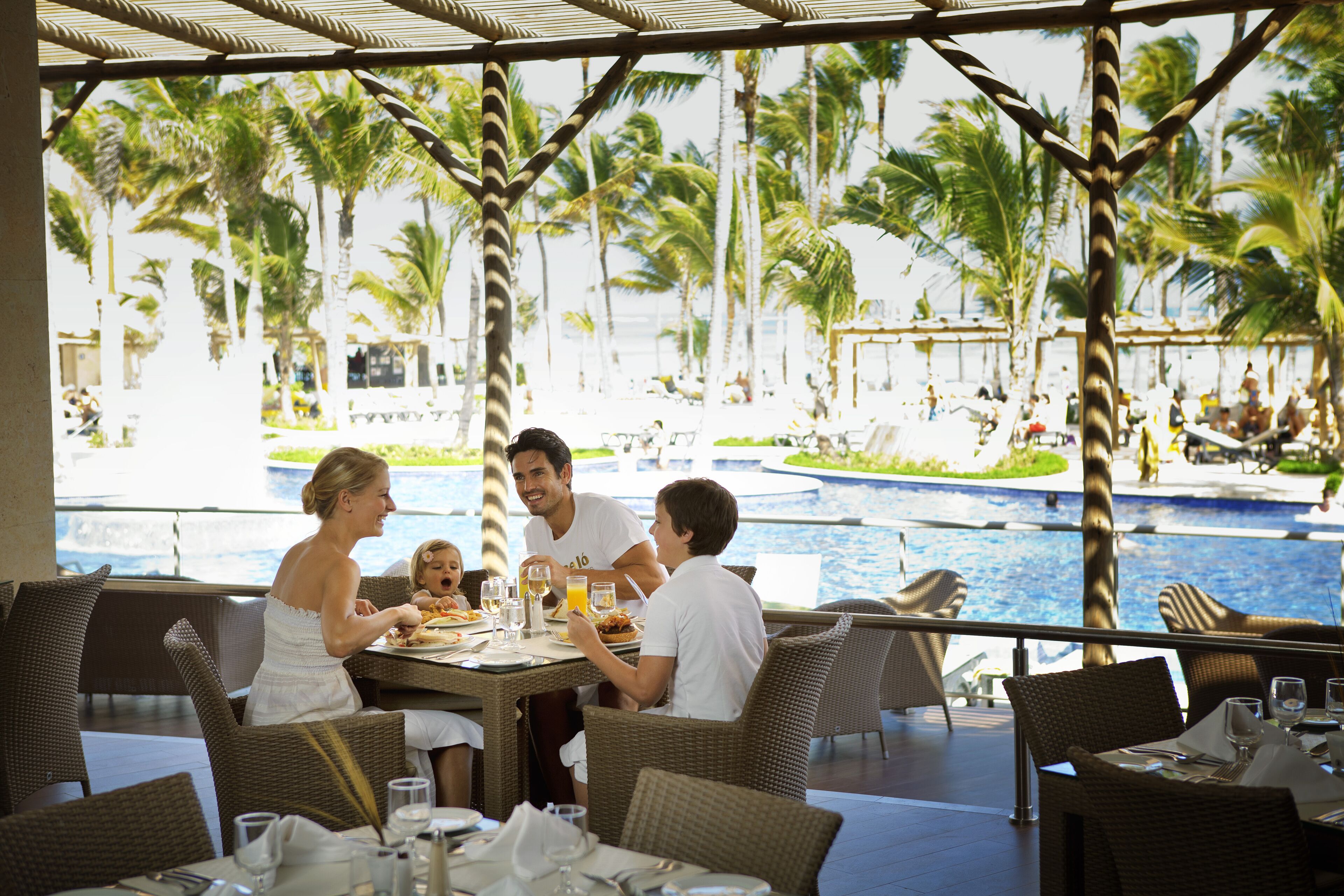 A dining area with wicker furniture, a pool view, and a tropical setting with palm trees.