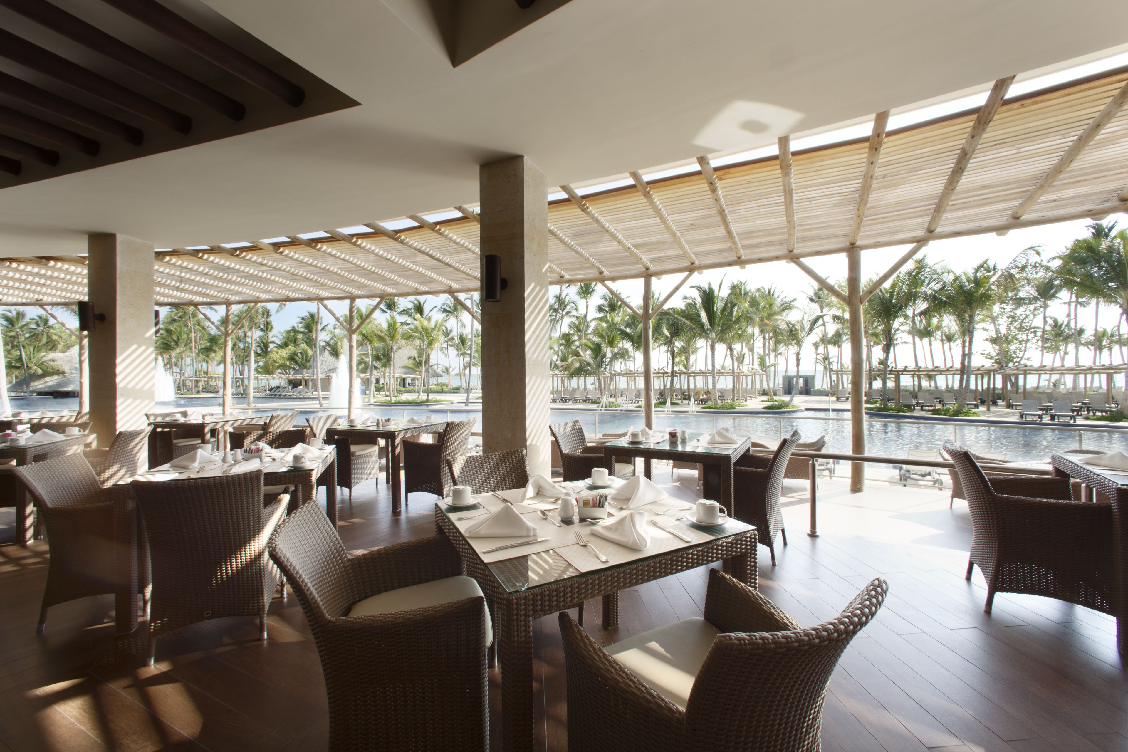 A restaurant with wicker furniture, a pool view, and a tropical setting.