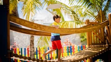 Un niño caminando sobre un puente de cuerda de madera en un entorno al aire libre, con palmeras y decoraciones coloridas.