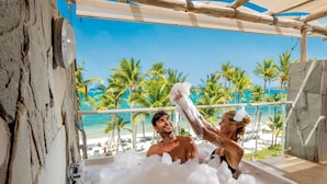 A bathtub filled with bubbles, a wooden structure, and a beach view with palm trees.