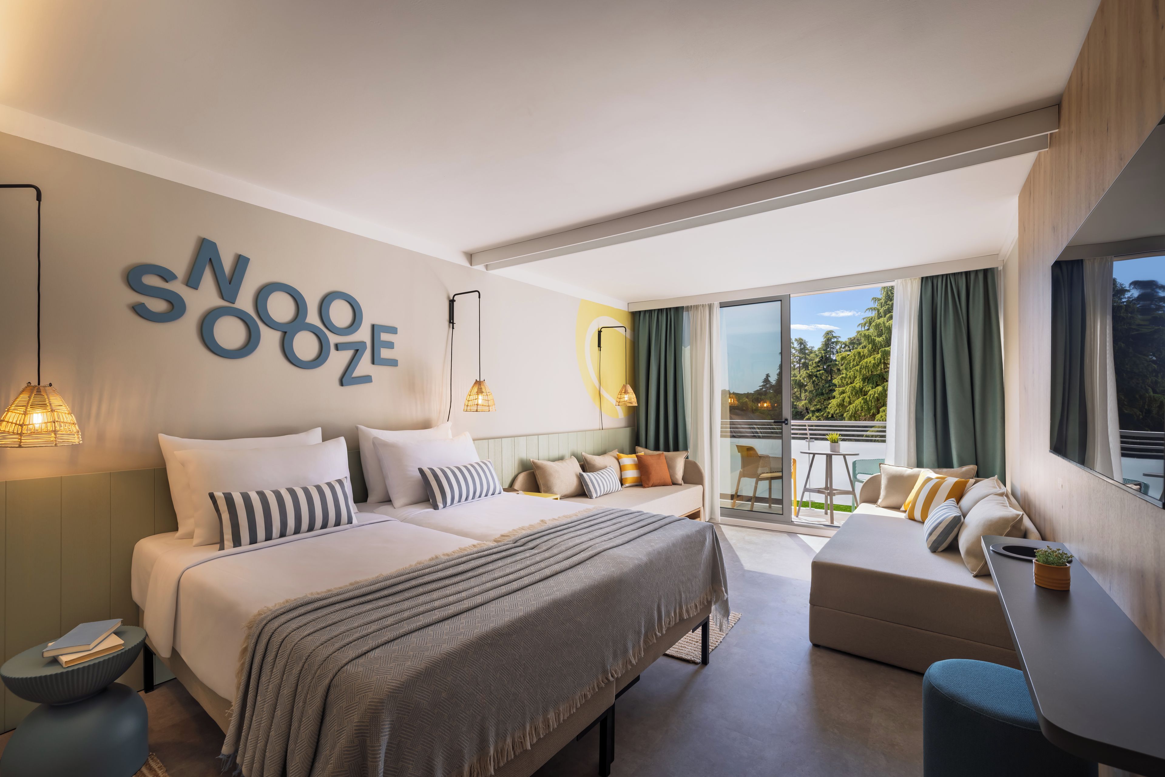A modern hotel room with a large bed, a sofa, a balcony with a view, and a wall with the text 'NOOZE'.