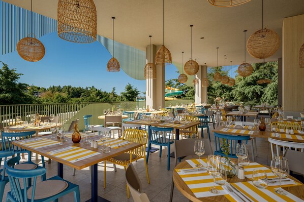 An outdoor dining area with tables and chairs, wicker hanging lights, and a view of trees and a pool. - Sunny Poreč by Valamar (Porec)