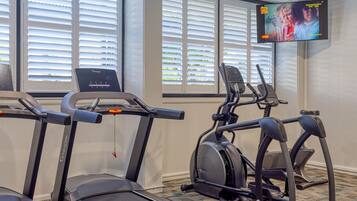 A fitness area with treadmills and an elliptical machine, a television mounted on the wall, and windows with blinds.