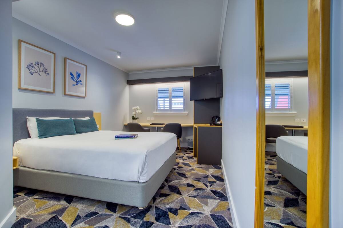A hotel room with a bed, desk, chair, TV, and two windows.