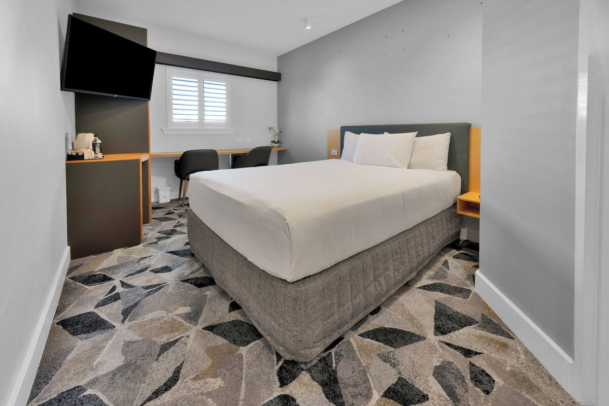 A hotel room with a large bed, a desk, a chair, a flat-screen TV, and a window with blinds.