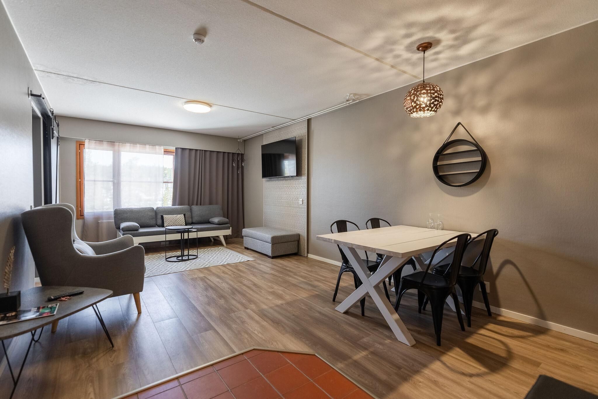 A modern living room with a sofa, a dining table with chairs, a TV mounted on the wall, and a patterned ceiling light.