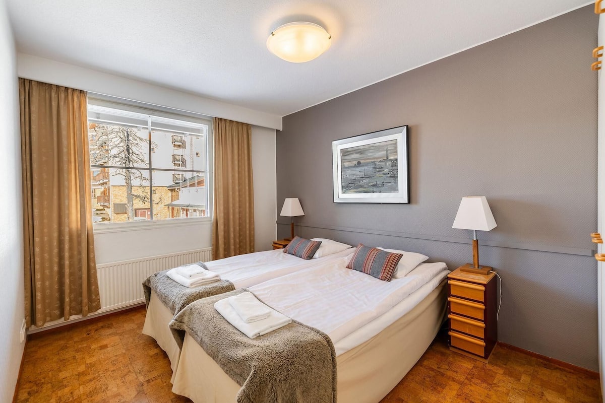 Une chambre d’hôtel avec un grand lit, deux tables de chevet, une fenêtre avec des rideaux, un tableau encadré au mur et un radiateur.