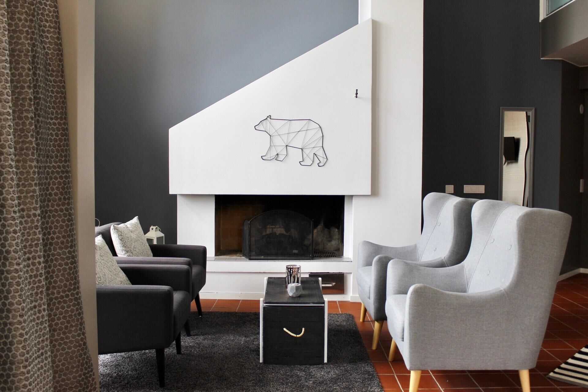 Een moderne woonkamer met een open haard, twee grijze fauteuils, een zwarte salontafel en een kamerkleed met een patroon.