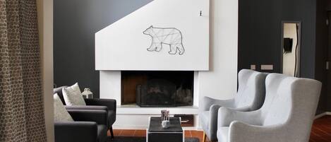 Een moderne woonkamer met een open haard, twee grijze fauteuils, een zwarte salontafel en een kamerkleed met een patroon.