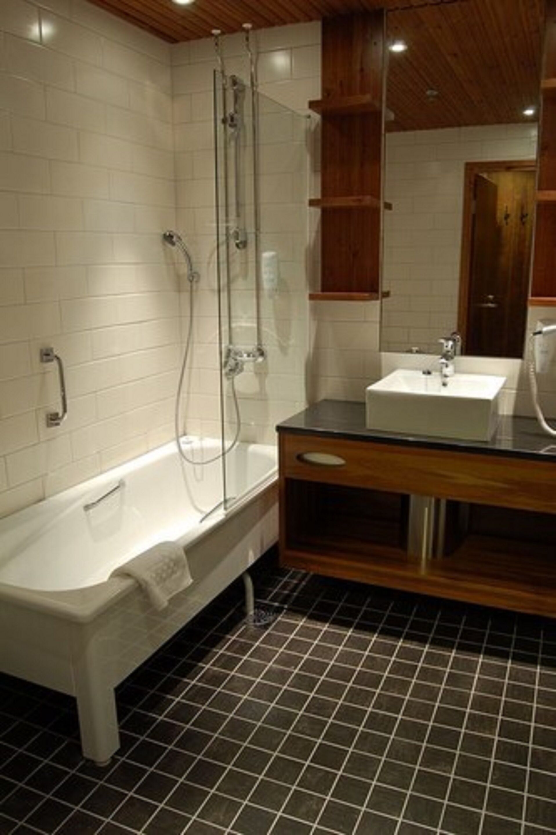 A modern bathroom with a white bathtub, a glass-enclosed shower, a wooden vanity with a sink, and a tiled floor.