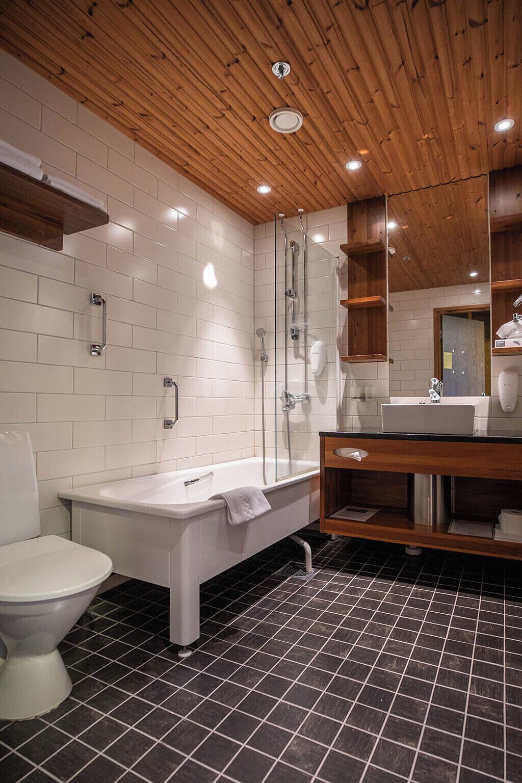 Un baño moderno con techo de madera, ducha con cerramiento de vidrio, bañera blanca y piso de baldosas negras.