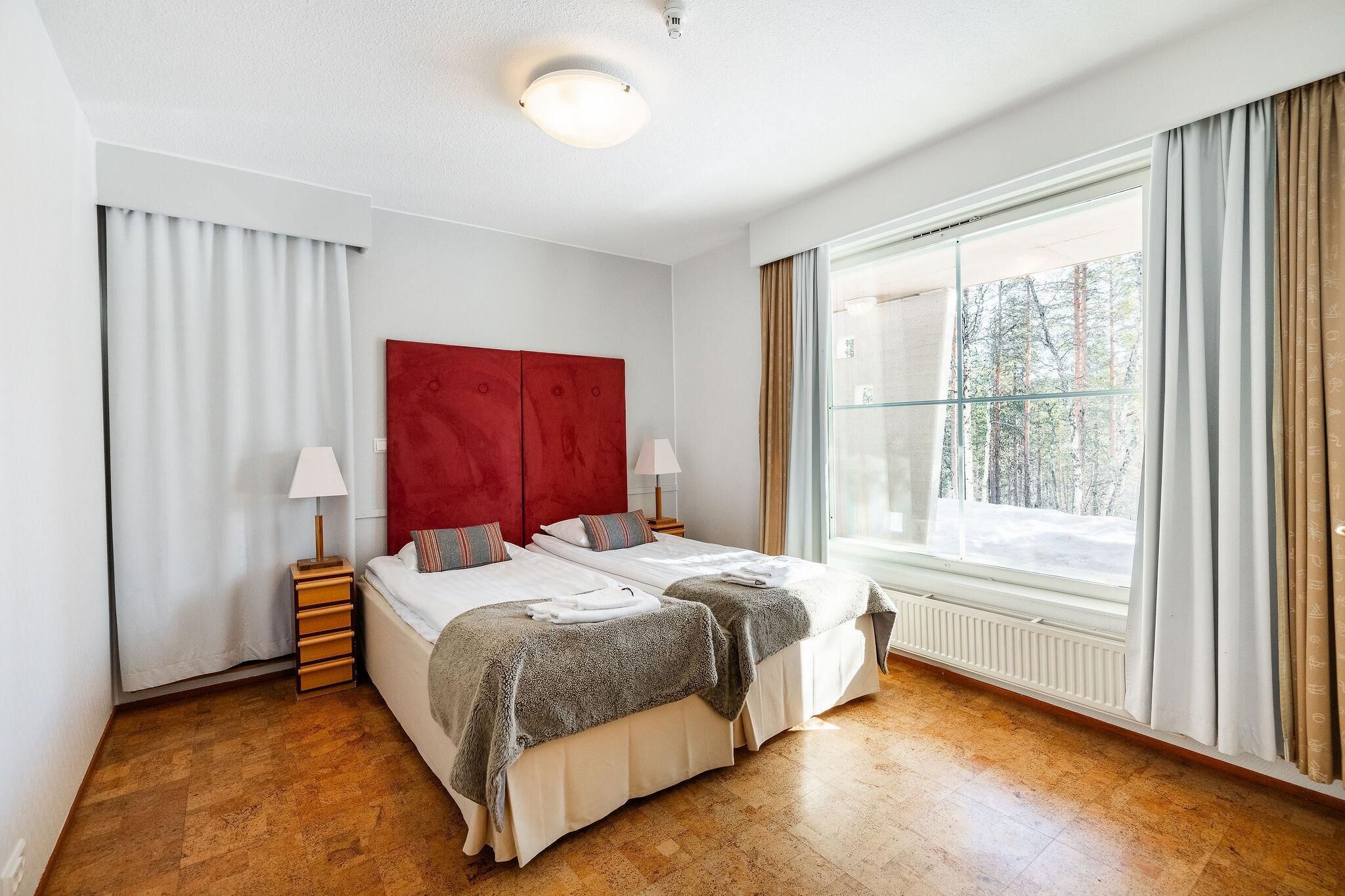 A hotel room with two beds, a red headboard, a window with curtains, and a wooden floor.