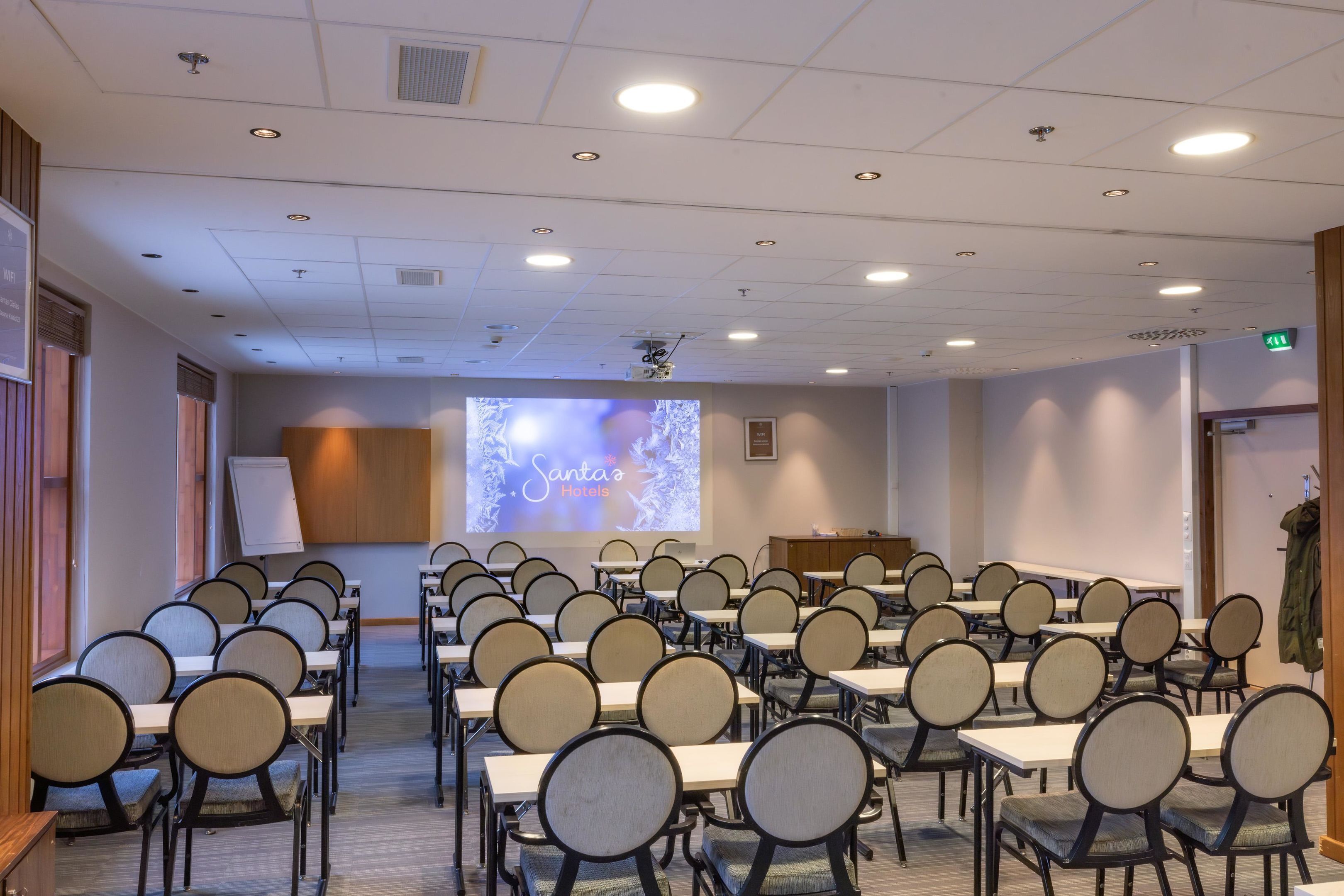 Un salón de conferencias con filas de mesas y sillas, una pantalla de proyección y un atril.