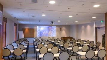 Un salón de conferencias con filas de mesas y sillas, una pantalla de proyección y un atril.