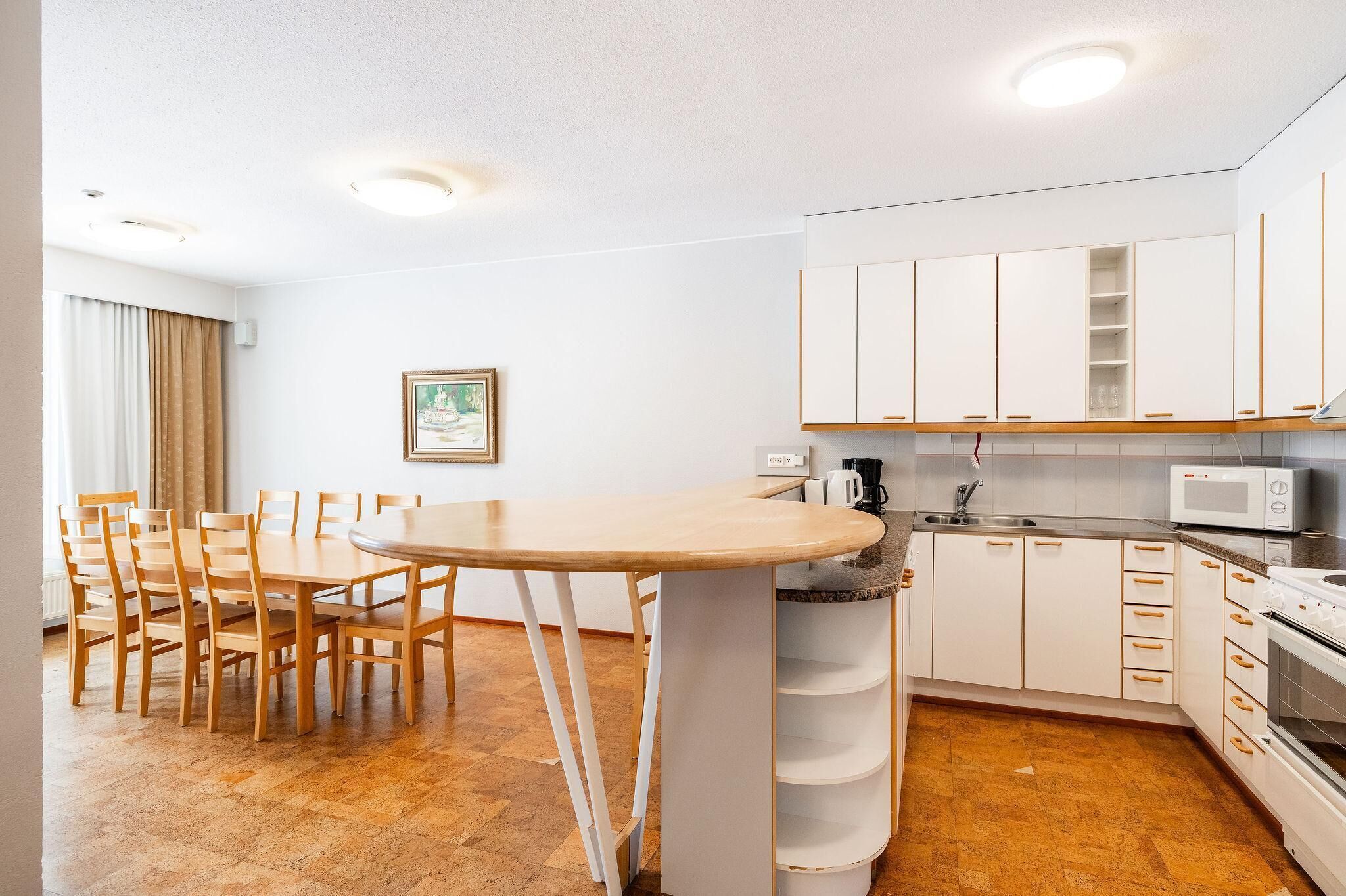 Een compacte keuken met witte kastjes, een magnetron en een fornuis. Er is een eettafel met stoelen en een lijst aan de muur.