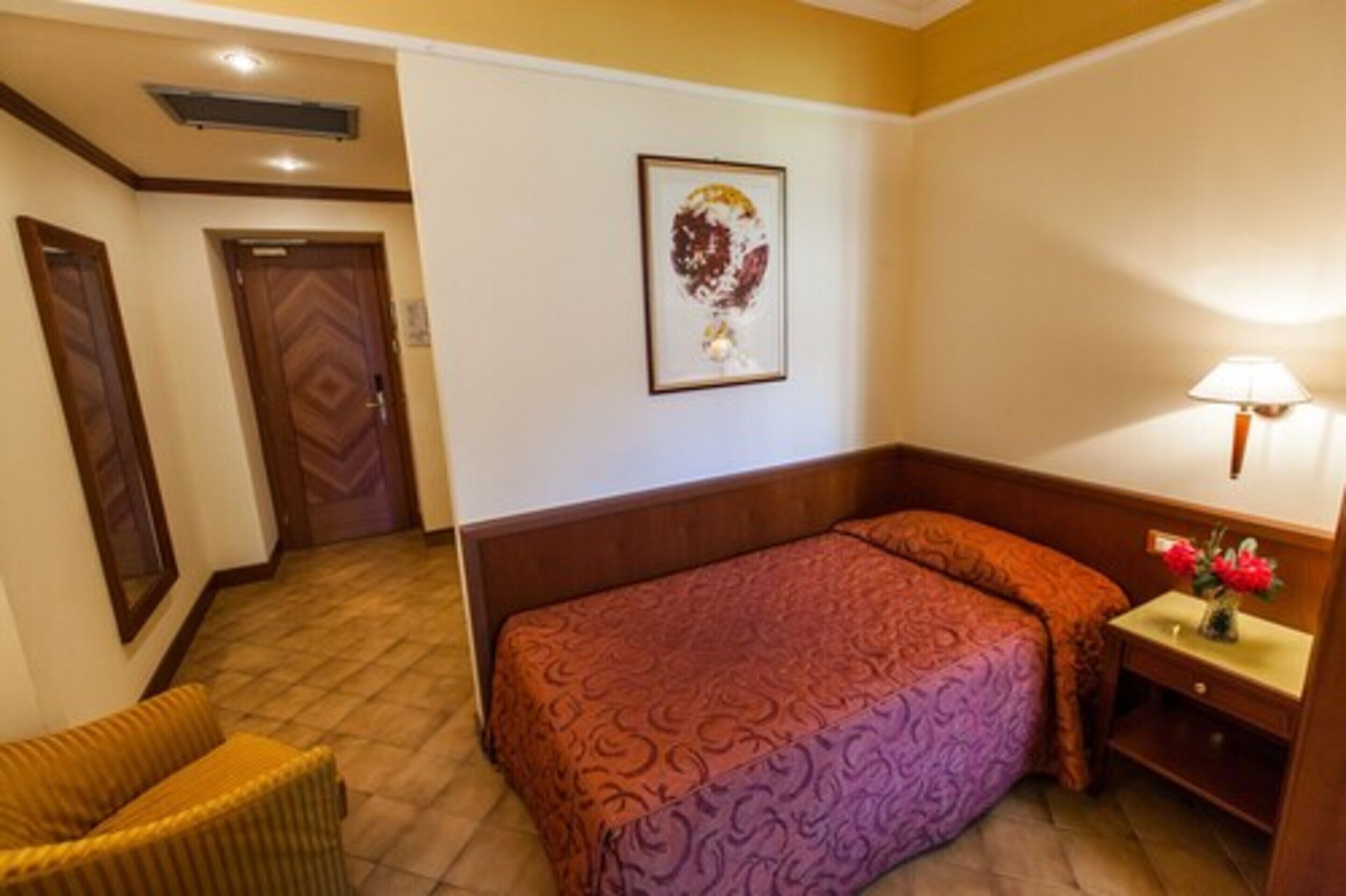 Une chambre d’hôtel avec un lit, une chaise, une table de chevet avec une lampe et un tableau encadré au mur.
