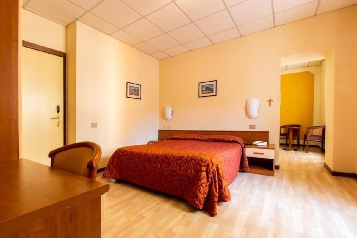 A hotel room with a wooden desk, a bed with a red bedspread, a nightstand, a chair, and a small table with a chair.