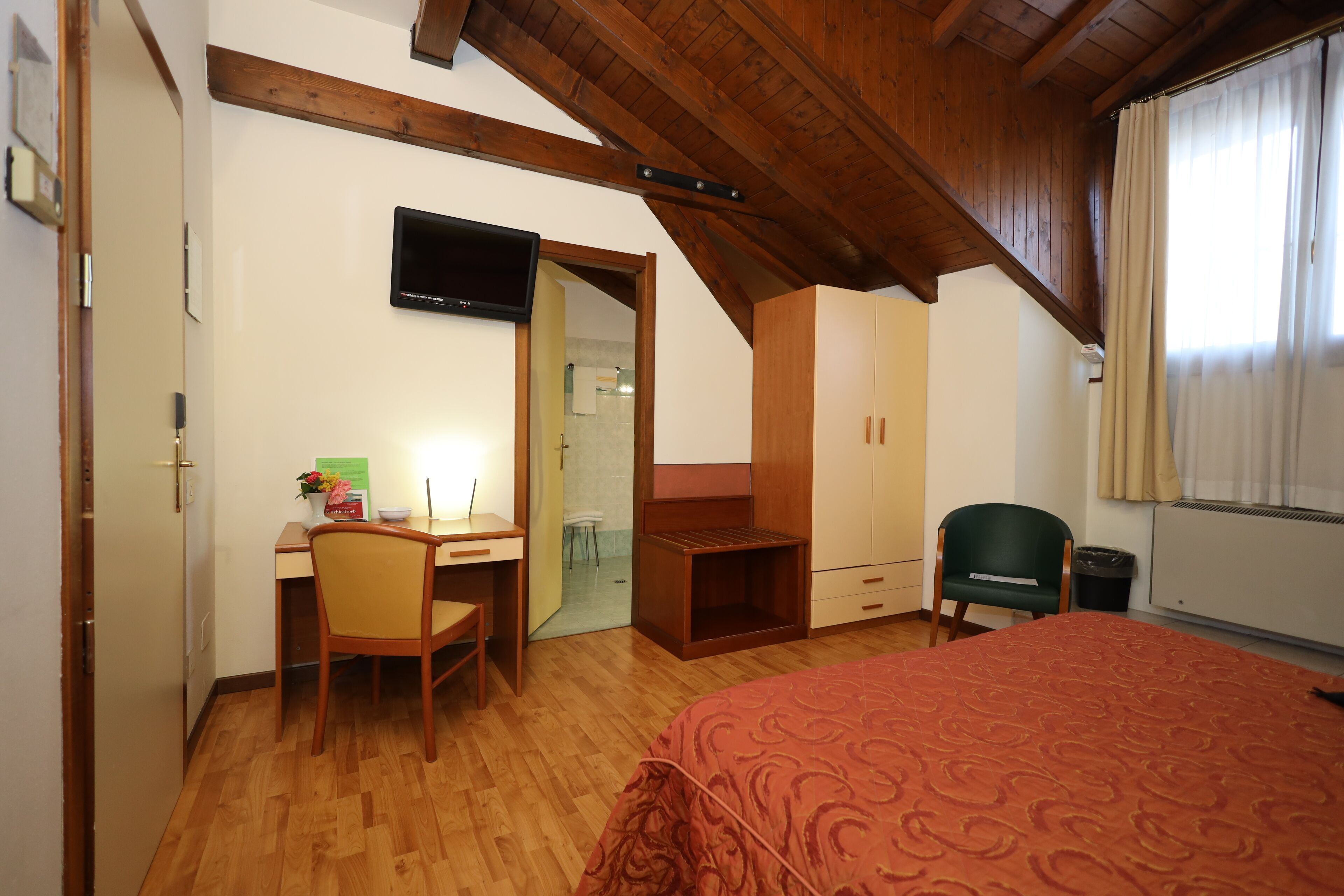 Ein Schlafzimmer mit Holzbalkendecke, einem Bett mit gemustertem Bettbezug, einem Schreibtisch mit Stuhl, einem Kleiderschrank und einem Fernseher an der Wand.