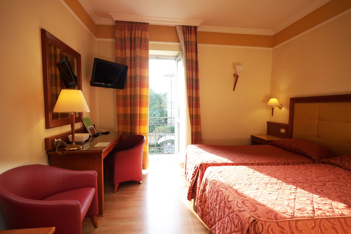 A hotel room with two beds, a desk with a lamp, a TV, and a chair.