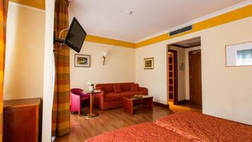 Une chambre d’hôtel comprenant un lit, un canapé, une petite table avec un vase, un téléviseur fixé au mur et une porte donnant sur une autre pièce.