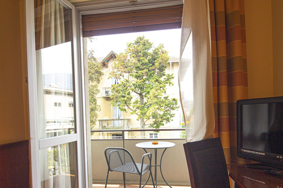 A hotel room with a bed, a TV, a chair, a table, and a balcony with a view of buildings and trees.