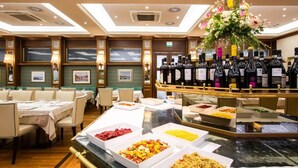 Een restaurantgedeelte in een hotel, met meerdere tafels, een buffet met diverse gerechten en een presentatie van wijn.