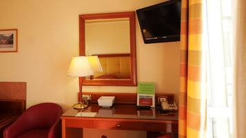 Ein Hotelzimmer mit einem Holztisch, einem roten Sessel, einem an der Wand befestigten Fernseher und einem Fenster mit Vorhängen.