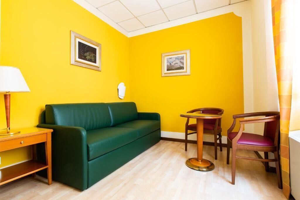 Una stanza con un divano verde, un tavolino di legno, una sedia e due quadri appesi alle pareti gialle.