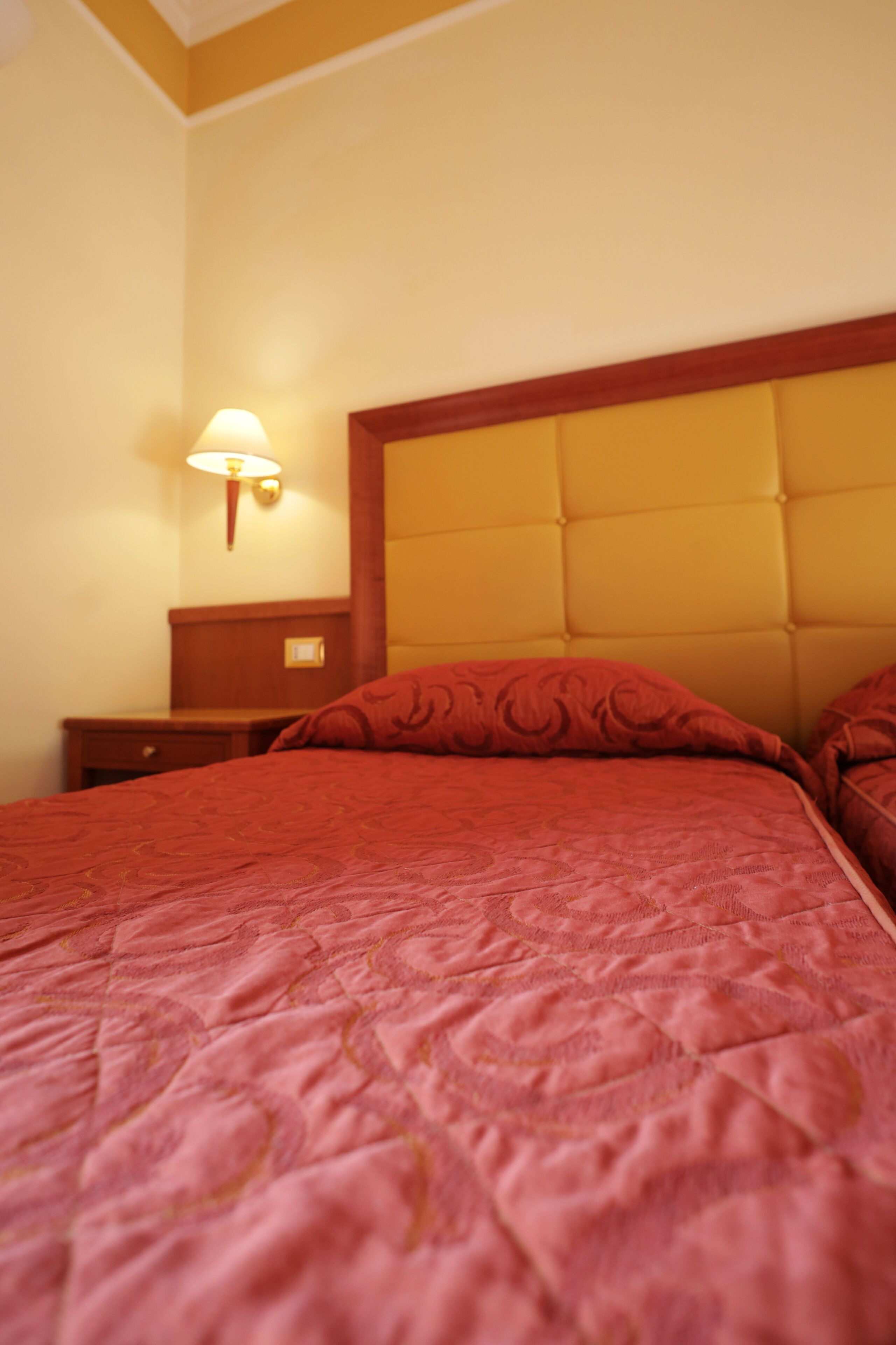 A hotel room with a large bed, a nightstand, and a wall-mounted lamp.