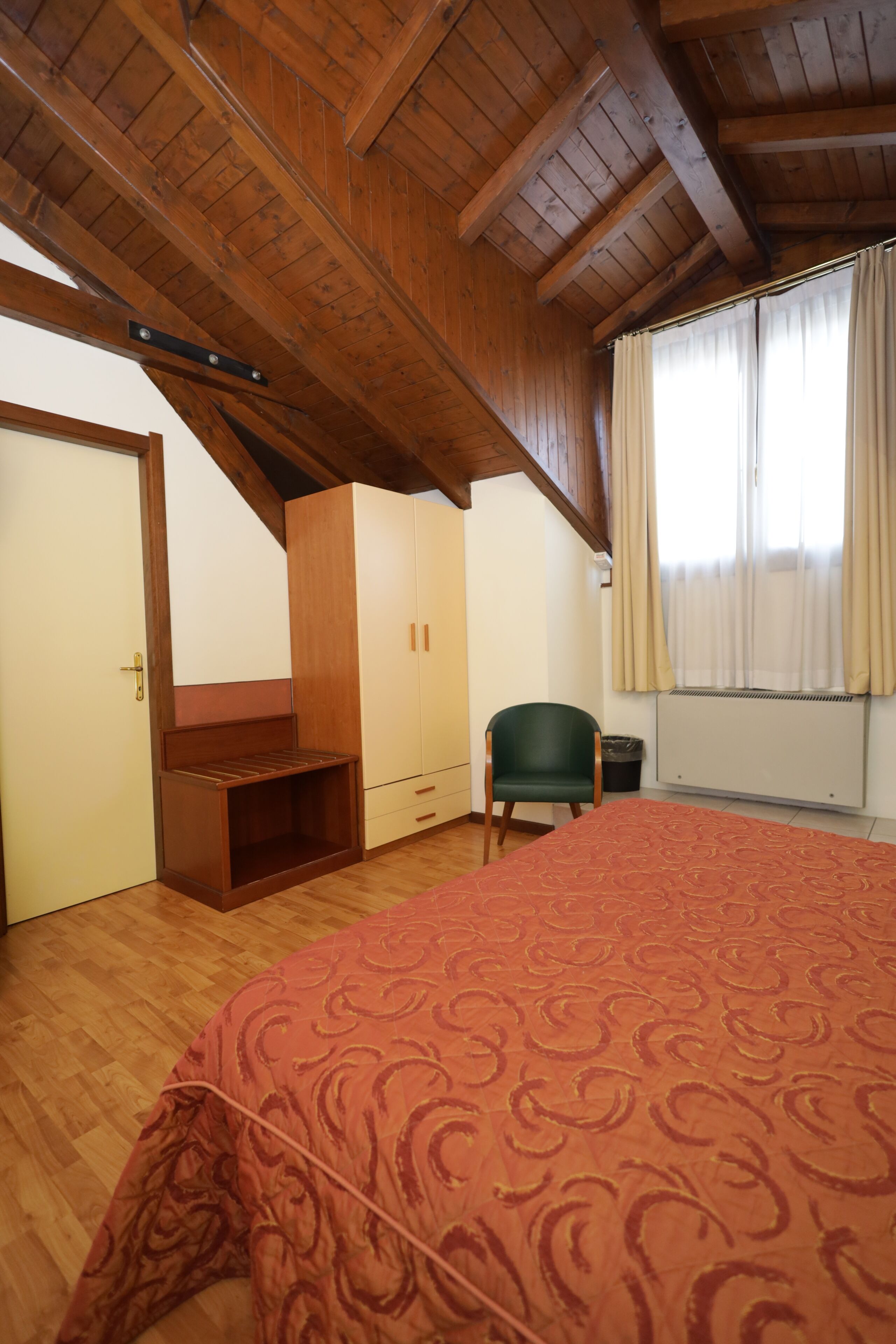 A bedroom with a wooden ceiling, a bed with a patterned bedspread, a green chair, and a radiator.