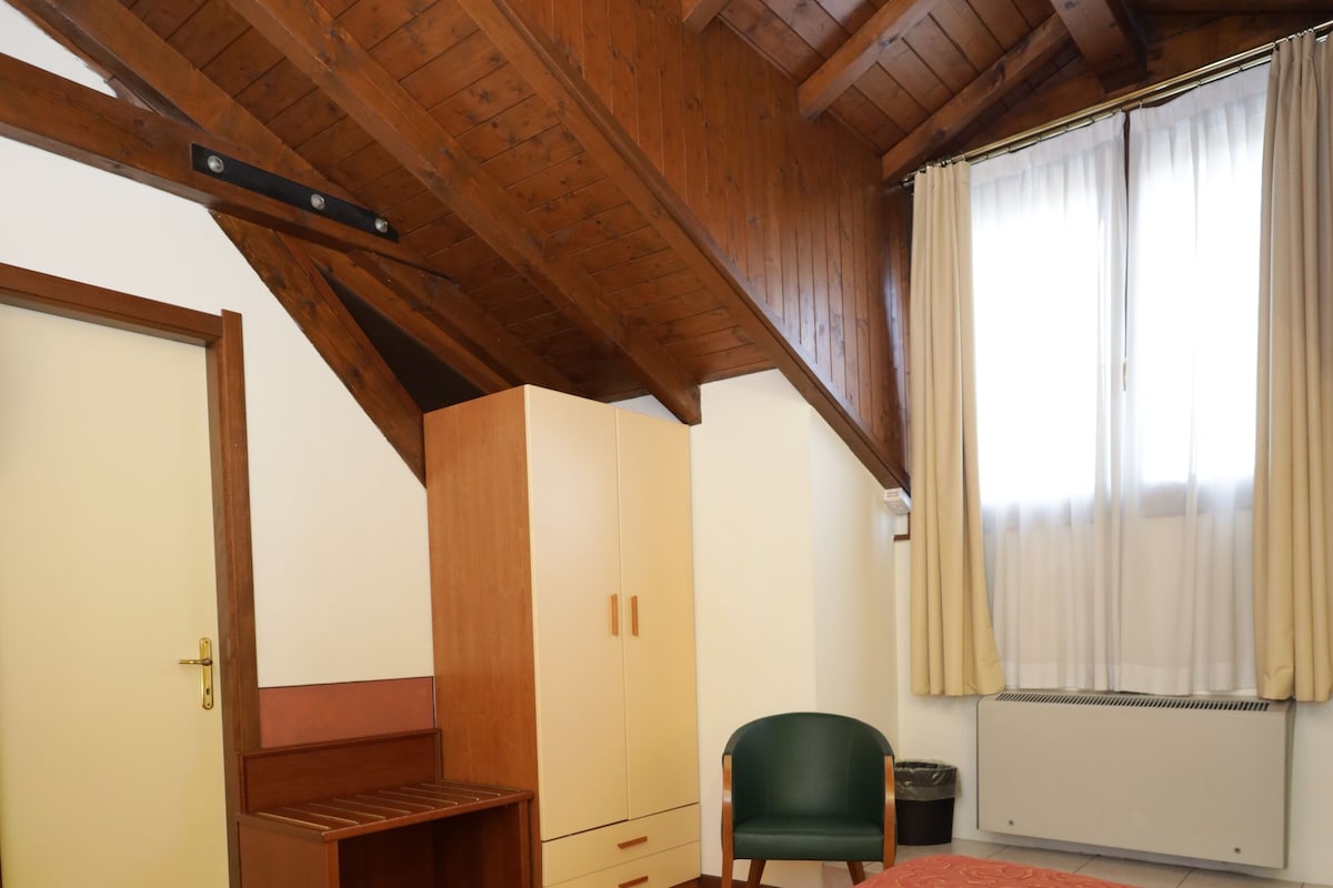 A bedroom with a wooden ceiling, a bed with a patterned bedspread, a green chair, and a radiator.