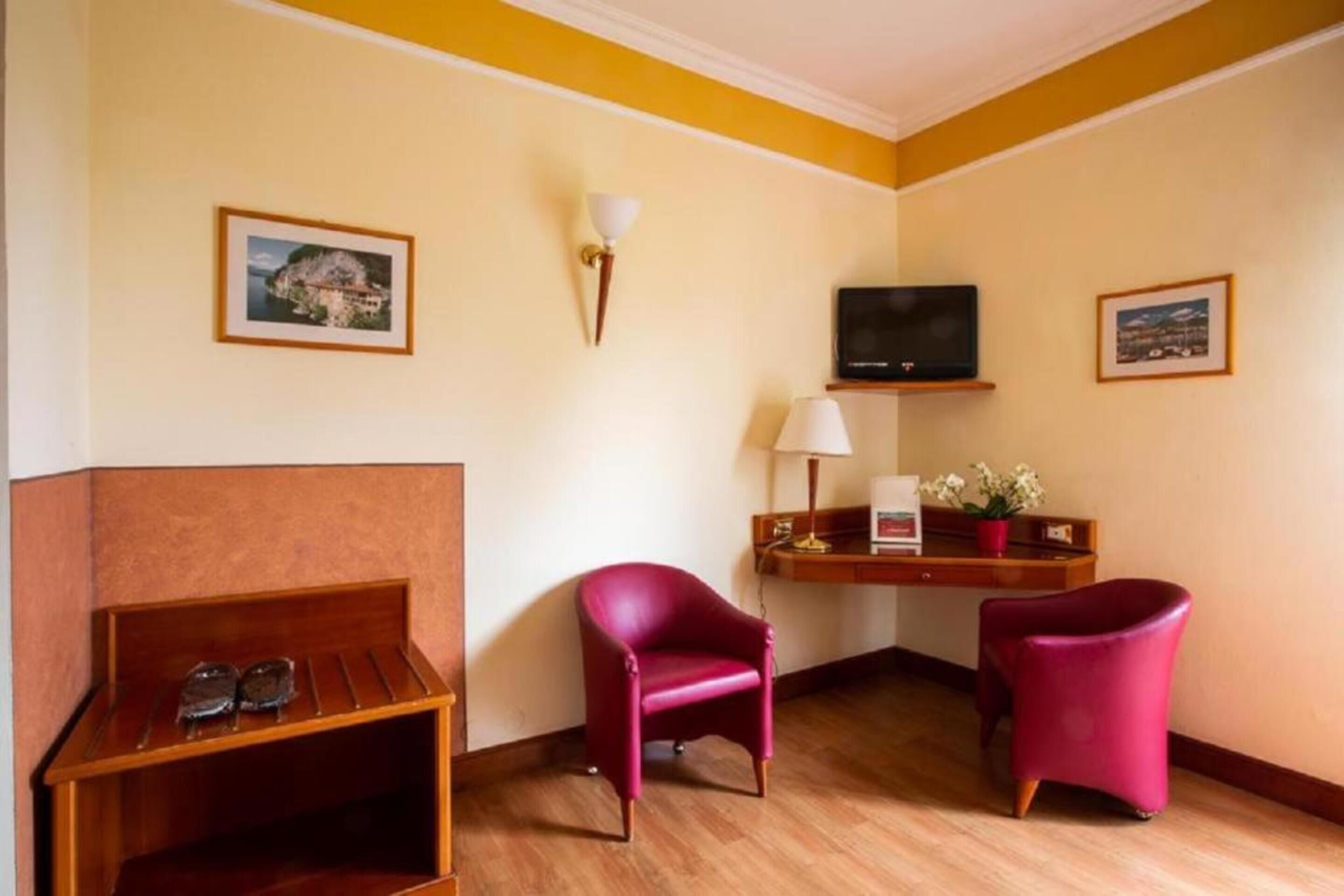 A hotel room with a wooden bench, two red chairs, a desk with a television, a lamp, and framed pictures on the walls.