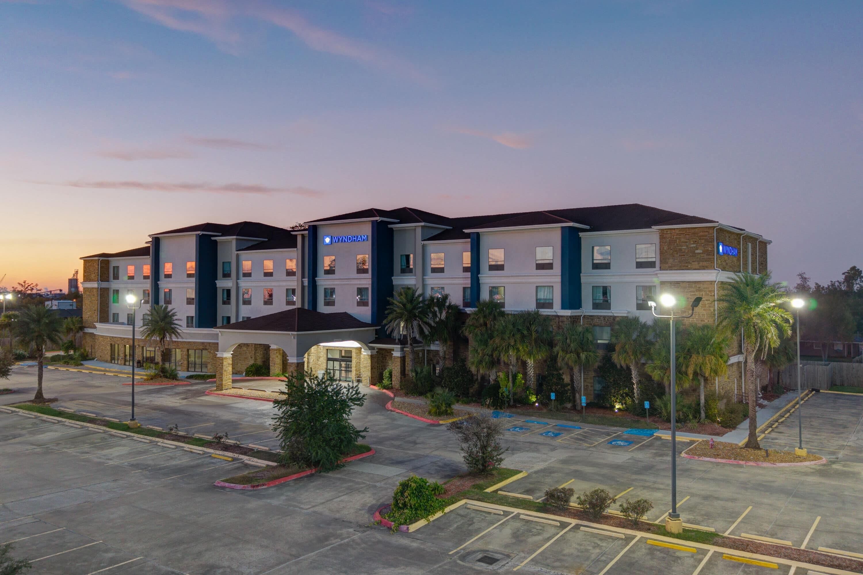 A multi-story hotel with a modern design, featuring a parking lot and landscaped areas with palm trees.
