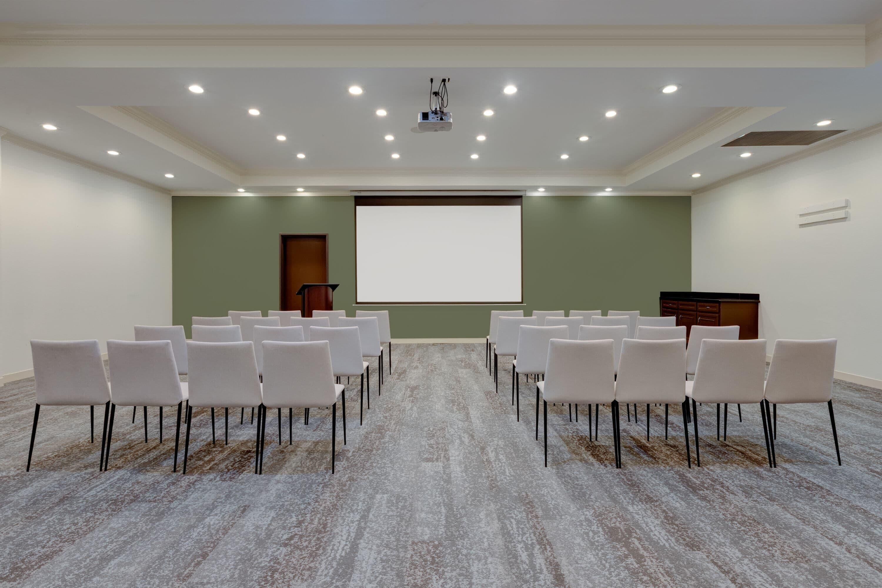 Ein Konferenzraum mit Reihen weißer Stühle, einer Projektionsleinwand und einem Teppichboden.